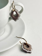 Versilana cocoa charm earring with a thin gold hoop resting on a neutral coffee cup and plate — a warm, elegant display highlighting the charm’s soft brown tone and minimalist design.