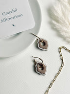 Versilana cocoa charm with a thin gold hoop displayed on a ceramic plate beside an affirmation card and a glimpse of a gold paperclip chain — a gentle reminder to shine with grace and confidence in every detail.