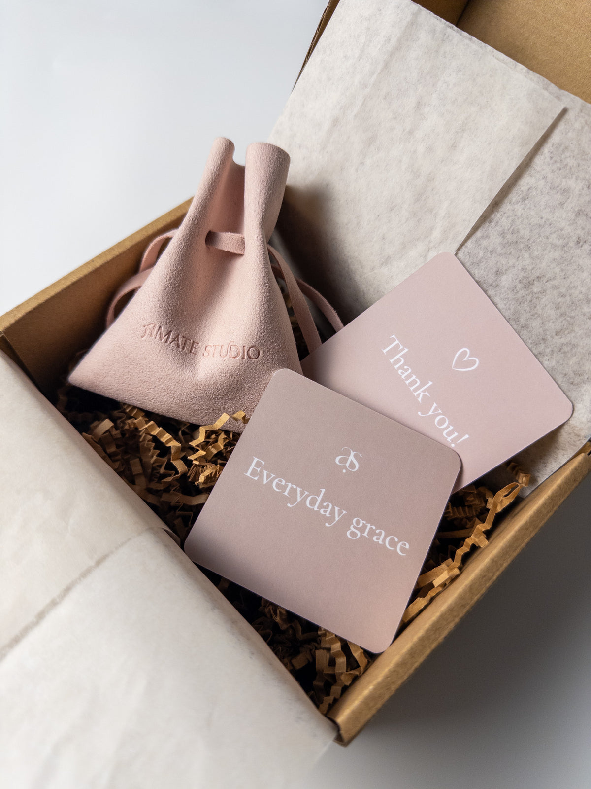Open Amate Studio shipping box with tissue paper, crinkle paper, jewelry pouch, thank you card, and Everyday Grace card included with jewelry orders.