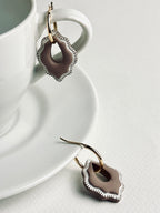 Versilana cocoa charm earring with a thin gold hoop resting on a neutral coffee cup and plate — a warm, elegant display highlighting the charm’s soft brown tone and minimalist design.