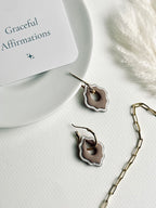 Versilana cocoa charm with a thin gold hoop displayed on a ceramic plate beside an affirmation card and a glimpse of a gold paperclip chain — a gentle reminder to shine with grace and confidence in every detail.