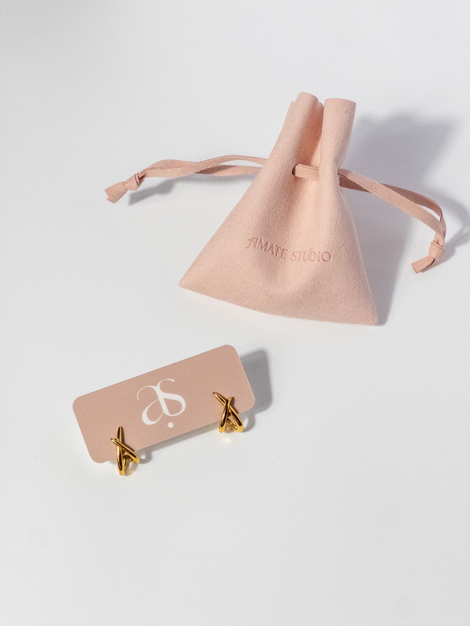 Lumen Studs gold crossover earrings displayed on a jewelry card beside a jewelry pouch.