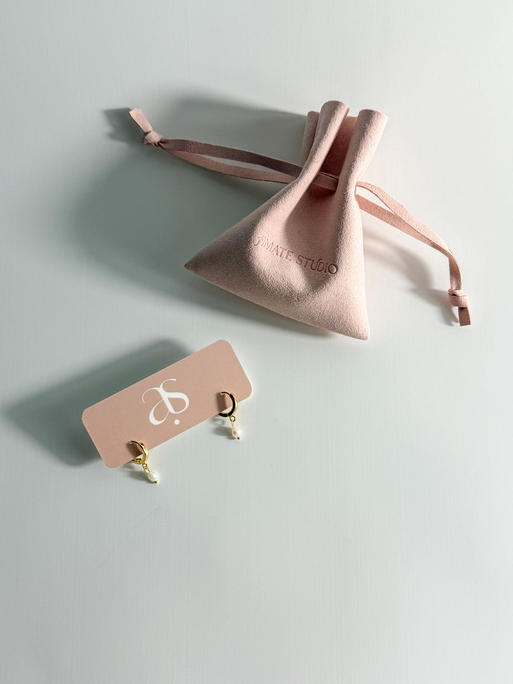 Meli gold-filled hoops with single freshwater pearls displayed on an Amate Studio earring card beside a branded microfiber pouch.