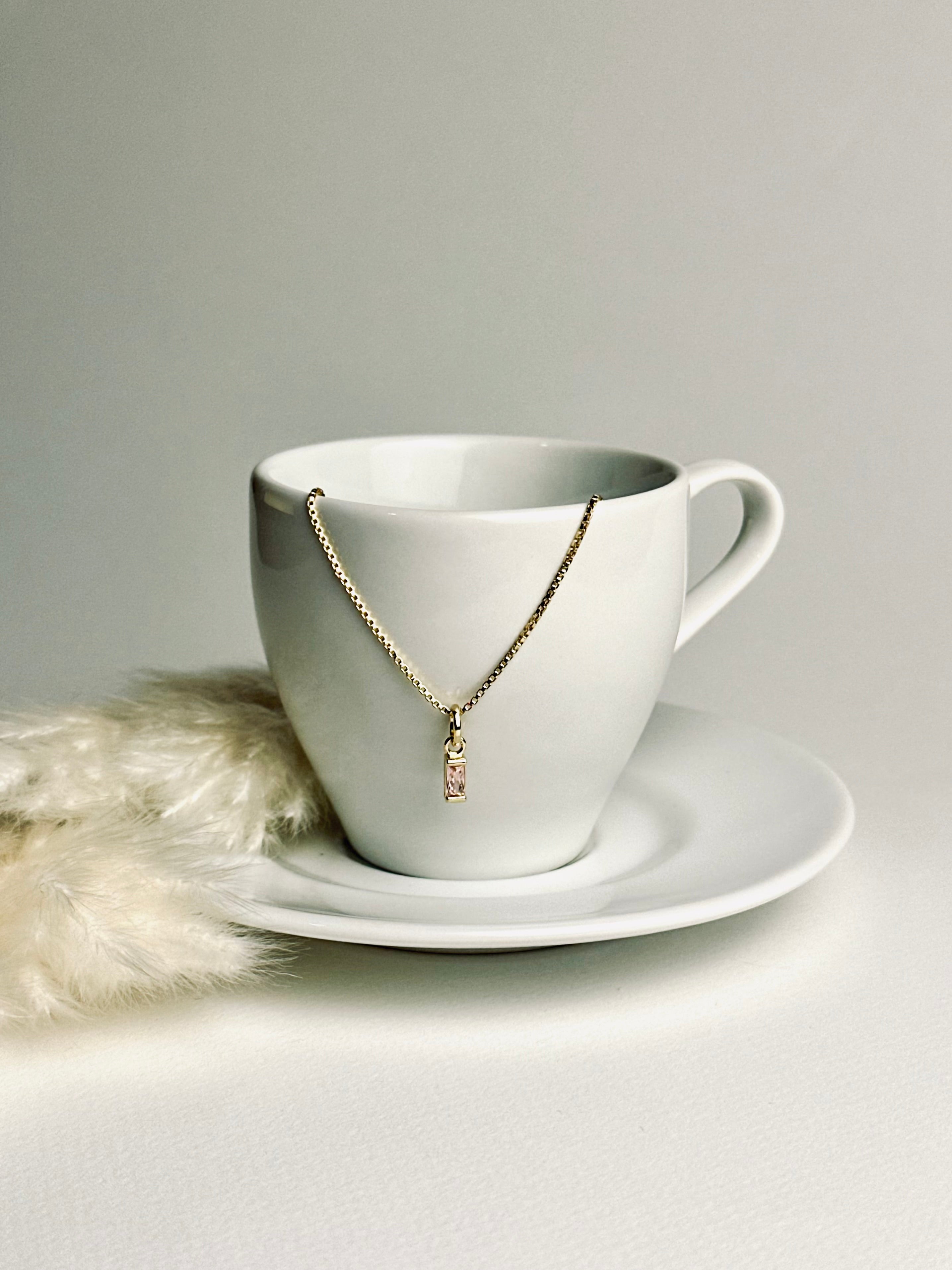 Aesthetic photo of a birthstone necklace delicately dangling over an espresso cup, with soft feather pampas in the background. The warm tones and gentle light evoke calm, elegance, and the meaningful beauty of everyday moments.