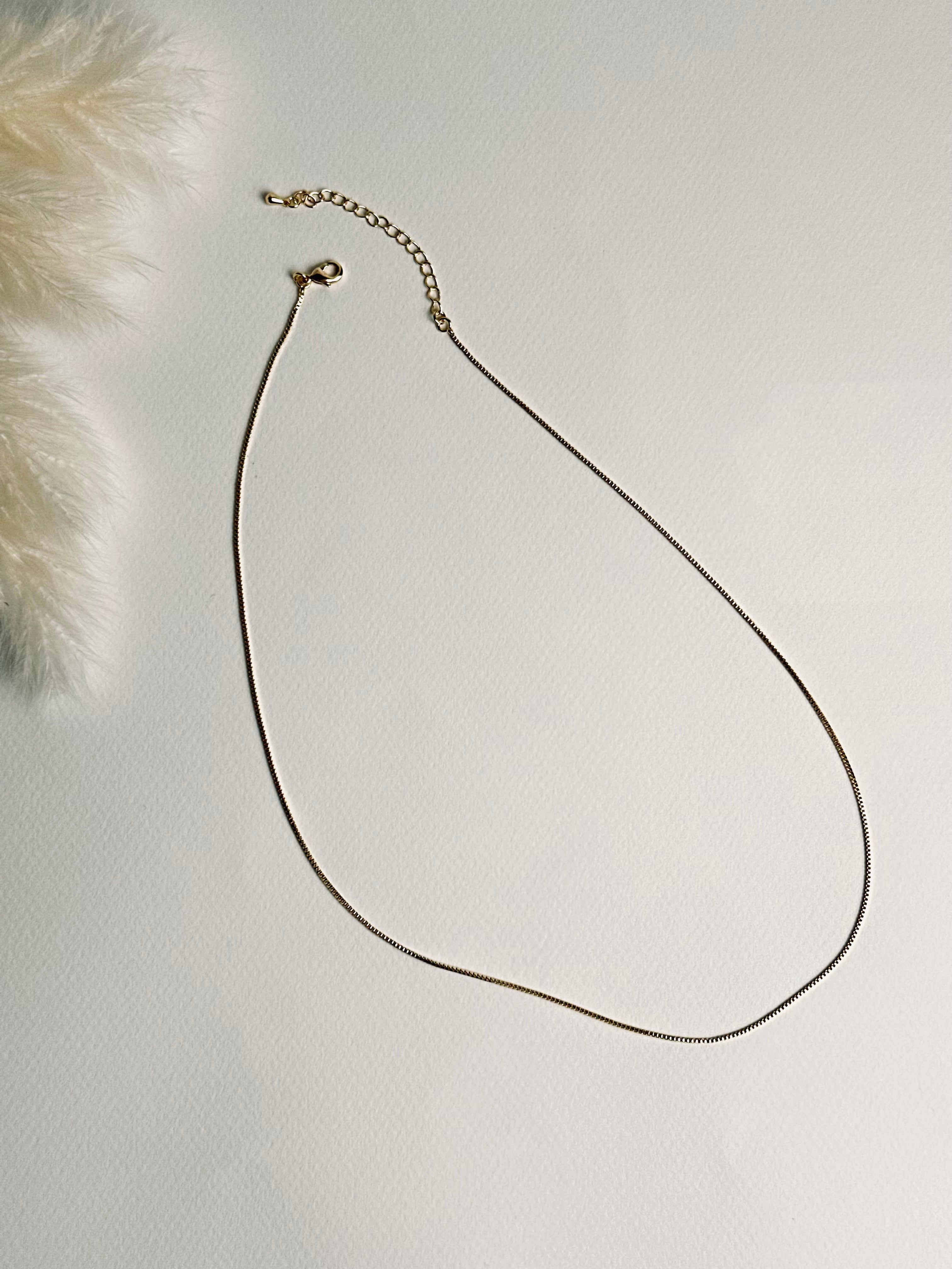 Serena — Gold Box Chain Necklace from the Timeless Grace collection, a thin gold-plated brass chain displayed on a book with focus on the lobster clasp and 2-inch extender. The delicate links and adjustable design highlight both elegance and practicality, symbolizing versatility, grace, and effortless style.