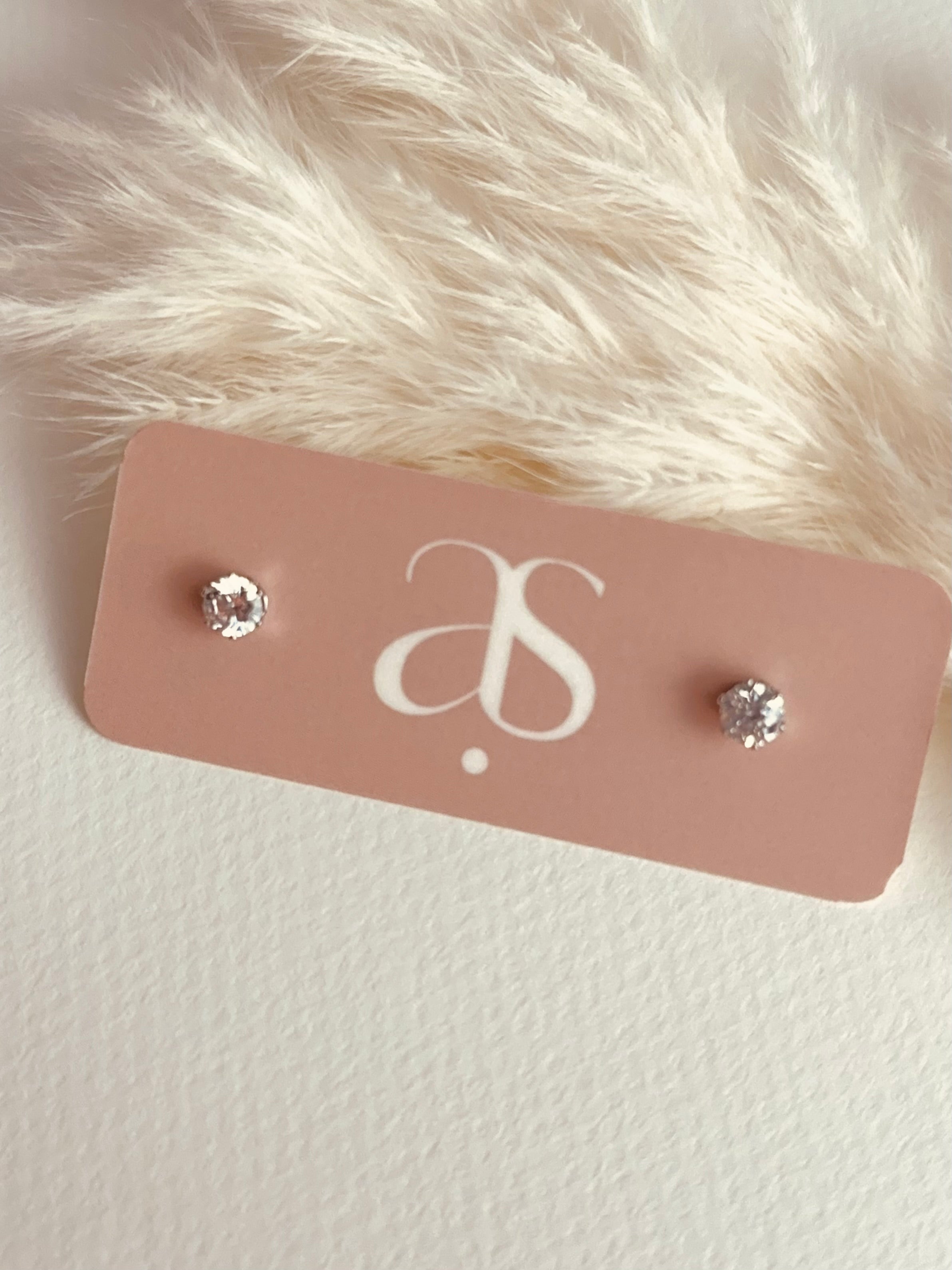 Daily Glow Studs – delicate 0.5mm stainless steel cubic zirconia earrings on blush Amate Studio card, styled with pampas feathers for a soft, elegant everyday grace look.
